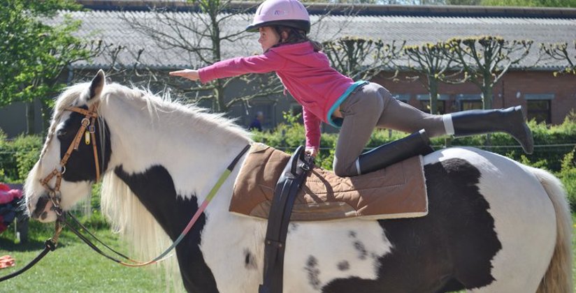 meisje doet een oefening met gestrekte arm en been op rug van een gevlekt paard